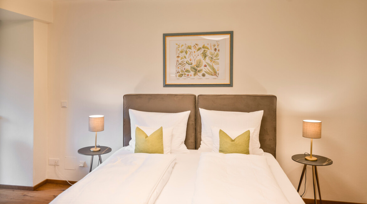 Elegant bedroom with twin beds, green accent pillows, bedside lamps, and nature-themed artwork above the headboard.