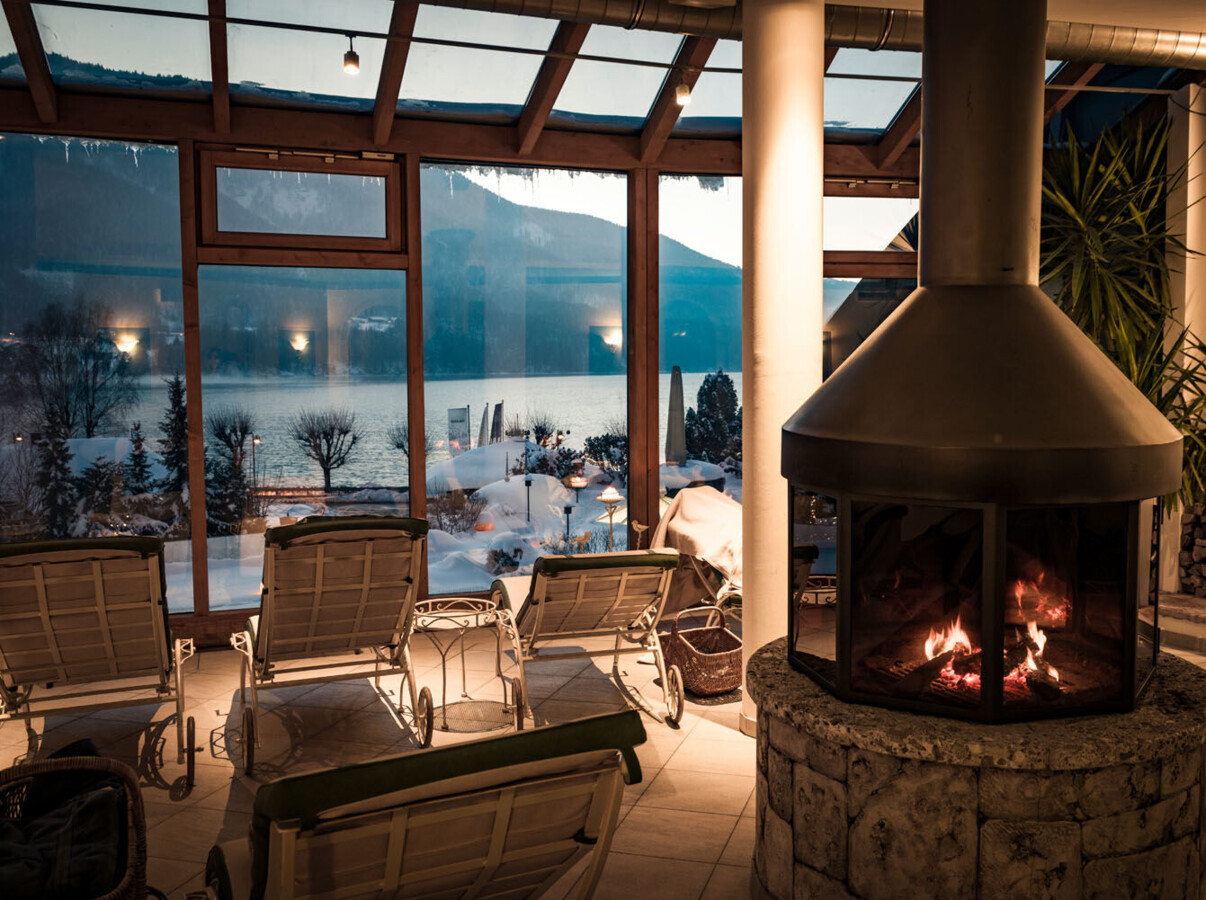 Cozy indoor seating with fireplace, overlooking Fuschlsee through large windows.