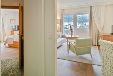 A cozy hotel suite with a bedroom and a living area, featuring elegant furnishings and a scenic view through large windows.
