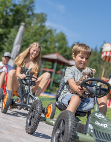Children riding pedal go-karts in an outdoor play area near a carousel, under a clear blue sky.