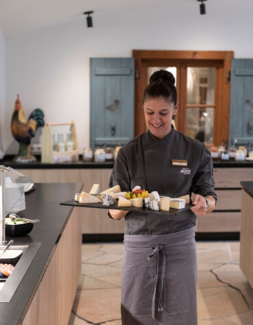 A staff member in a restaurant presents a cheese platter with various cheeses in a well-lit dining area.