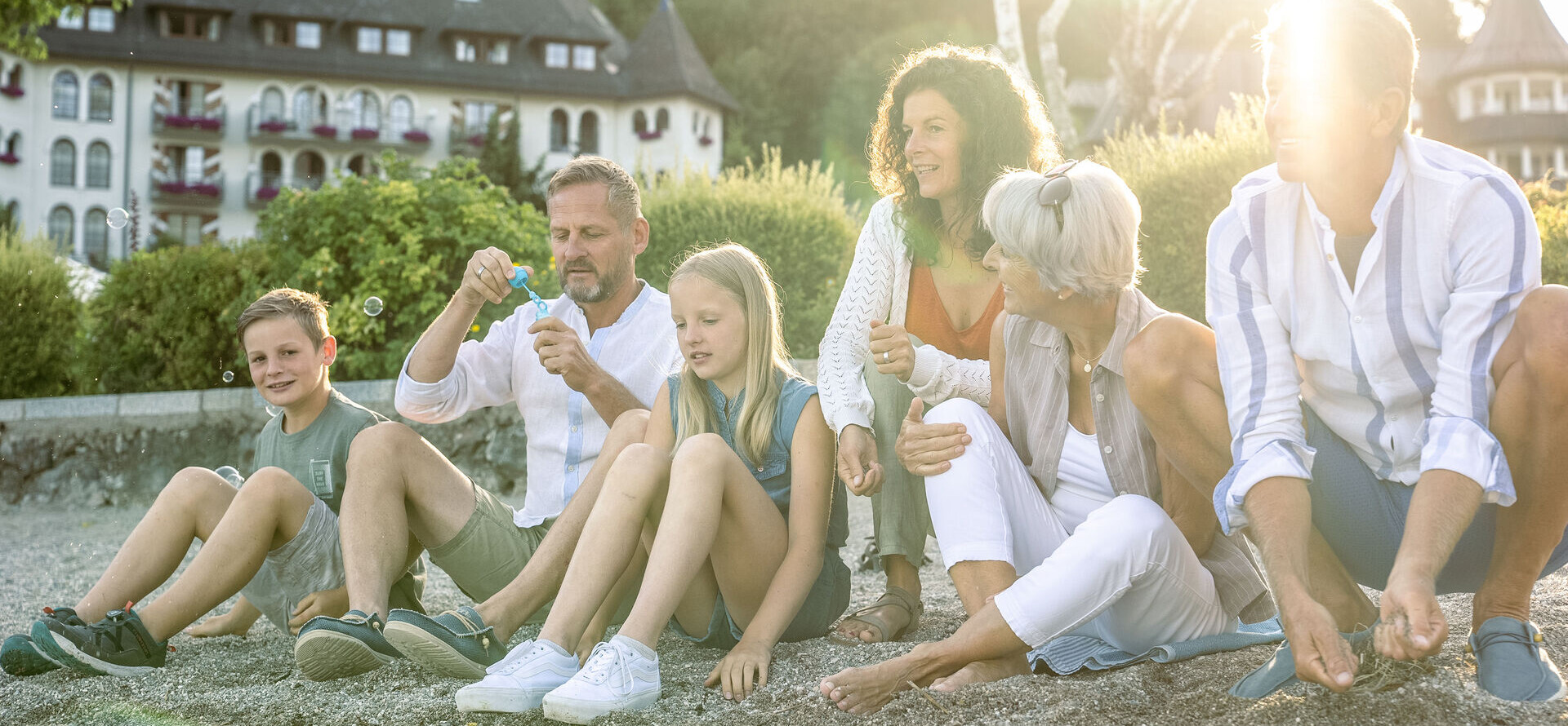A family enjoying a sunny day by the water, with a scenic hotel and lush greenery in the background.