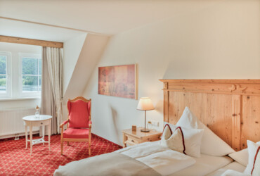 Gemütliches Hotelzimmer mit Holzbetten, rotem Sessel, Tisch und Lampe. Fenster mit Blick auf die Landschaft.