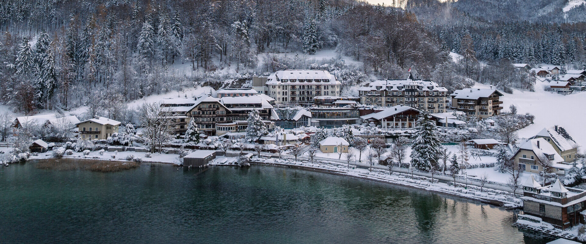 Winterlandschaft mit verschneiten Gebäuden am Fuschlsee, umgeben von bewaldeten Bergen bei Sonnenuntergang.