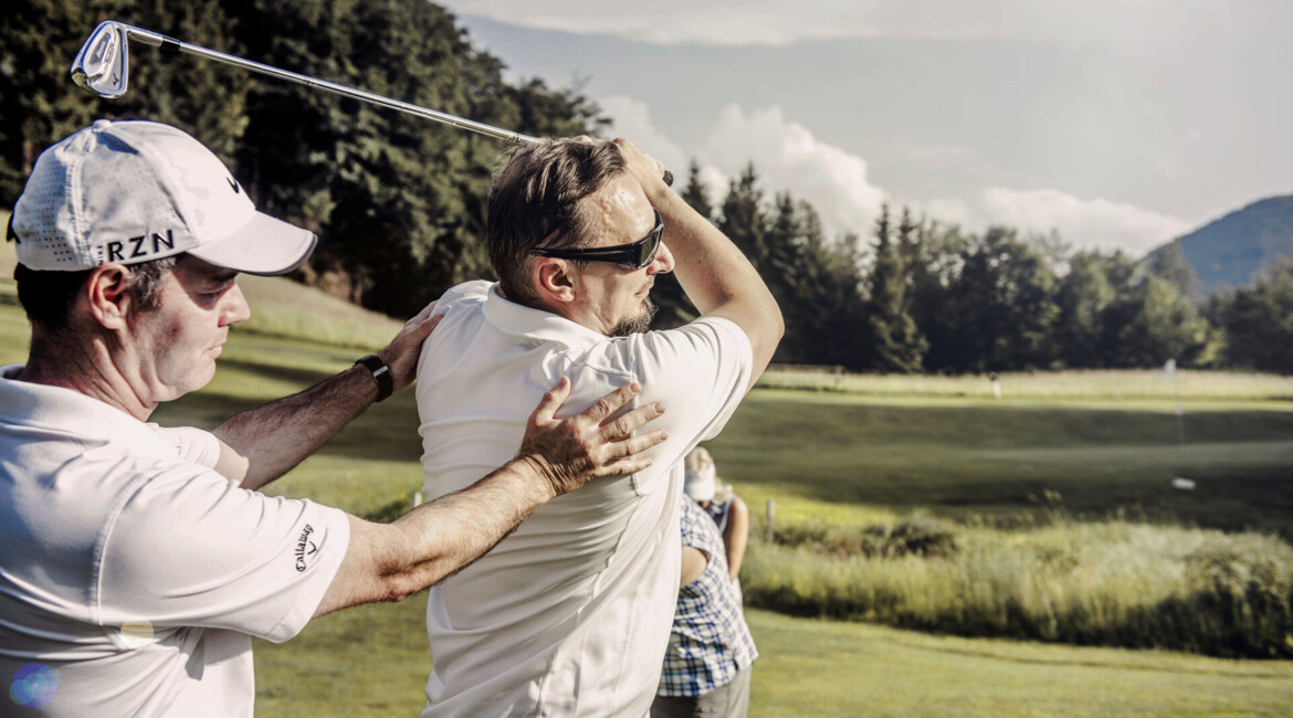 Zwei Männer spielen Golf auf einem grünen Feld, einer korrigiert den Schwung des anderen. Im Hintergrund sind Bäume und Berge zu sehen.