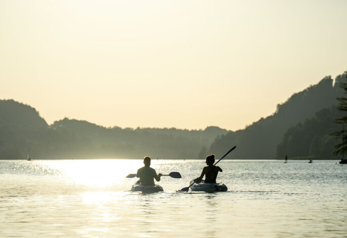 Zwei Personen paddeln im Sonnenuntergang auf einem ruhigen See, umgeben von Hügeln.