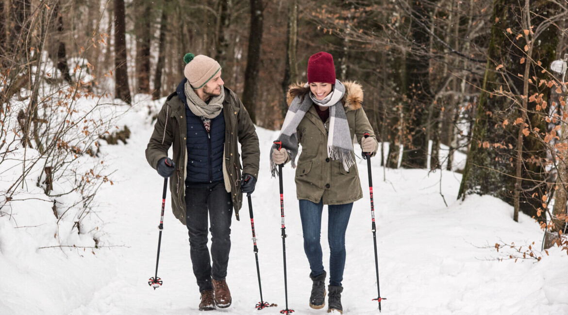 A couple enjoys a winter hike through a snowy forest, using trekking poles for support.