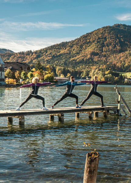 Drei Personen machen Yoga auf einem Steg am Fuschlsee, umgeben von herbstlichen Bergen und klarem Wasser.