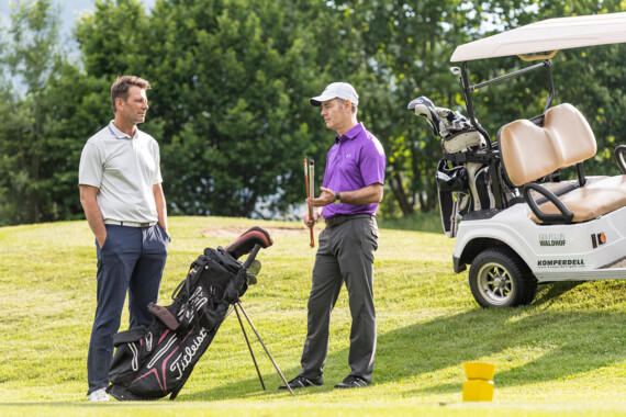 Two men talking on a golf course near a cart, one holding a club.