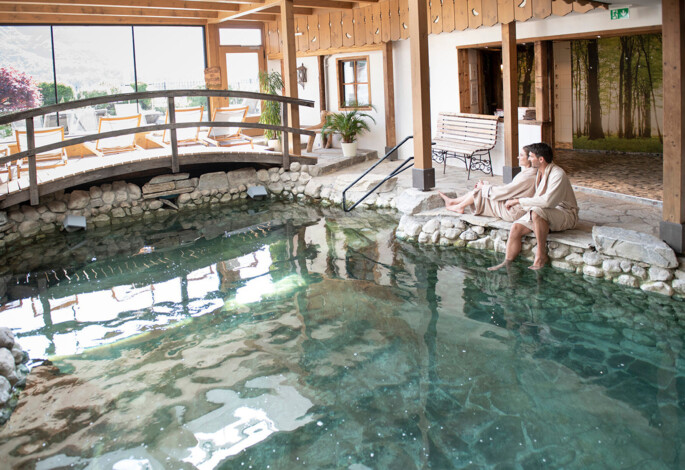 Zwei Personen entspannen am Rand eines Innenpools mit Steinverkleidung in einem gemütlichen Spa-Bereich.