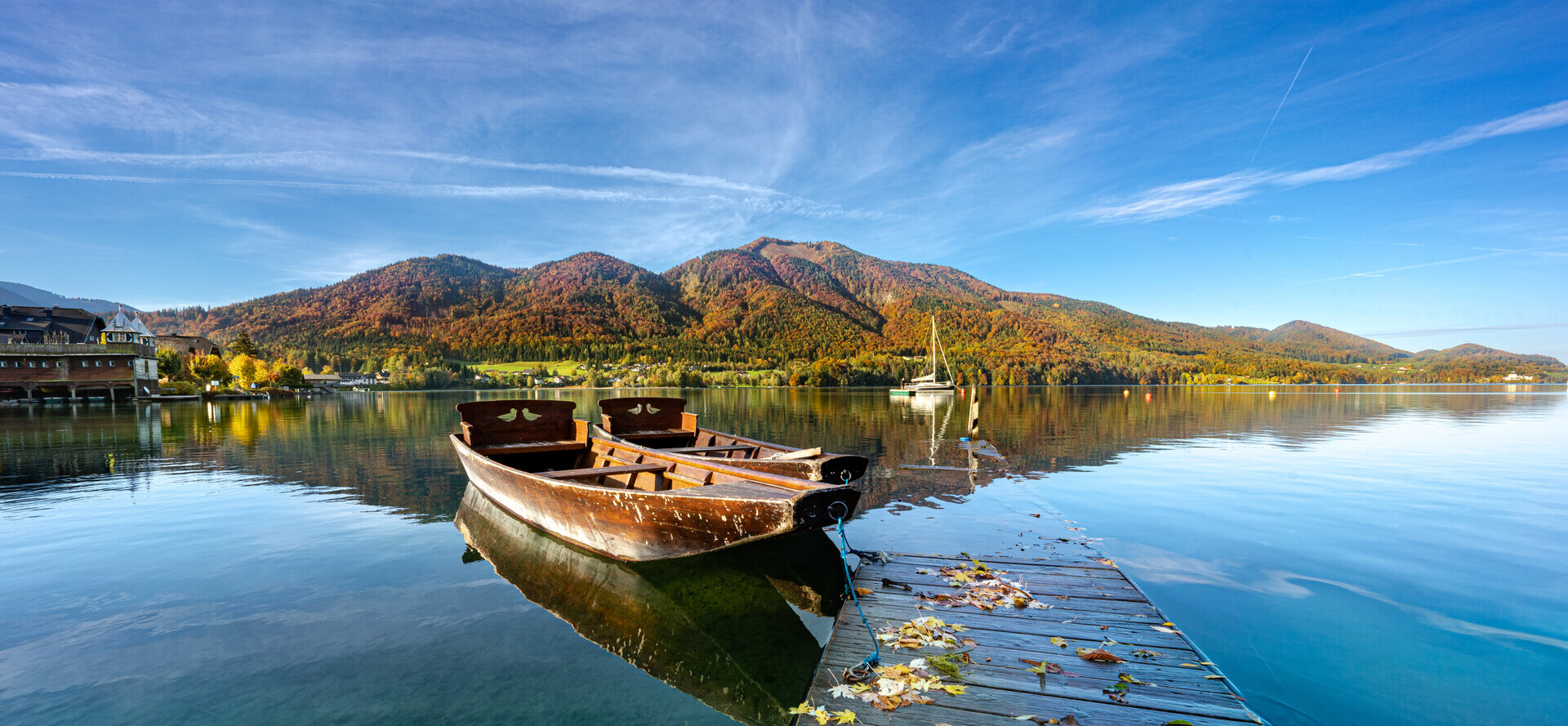 Ein Holzboot liegt an einem Steg auf einem ruhigen See, umgeben von herbstlichen Bergen unter klarem blauem Himmel.