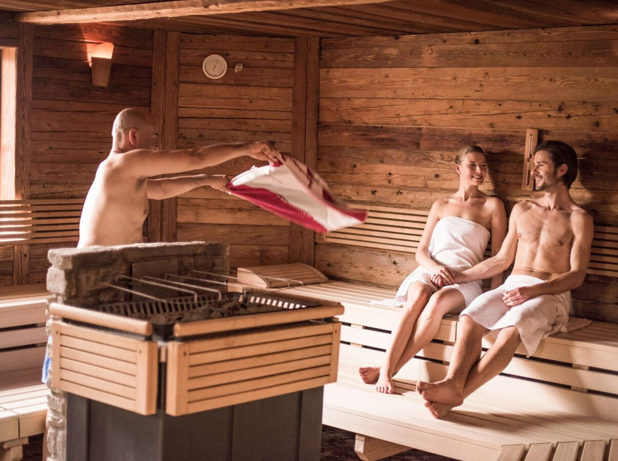 Personen entspannen in einer Sauna mit Holzwänden, während ein Mann ein Tuch wedelt, um die Hitze zu verteilen.
