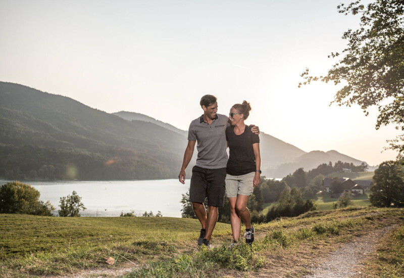 Ein Paar spaziert bei Sonnenuntergang am Fuschlsee entlang, umgeben von grünen Hügeln und ruhiger Natur.