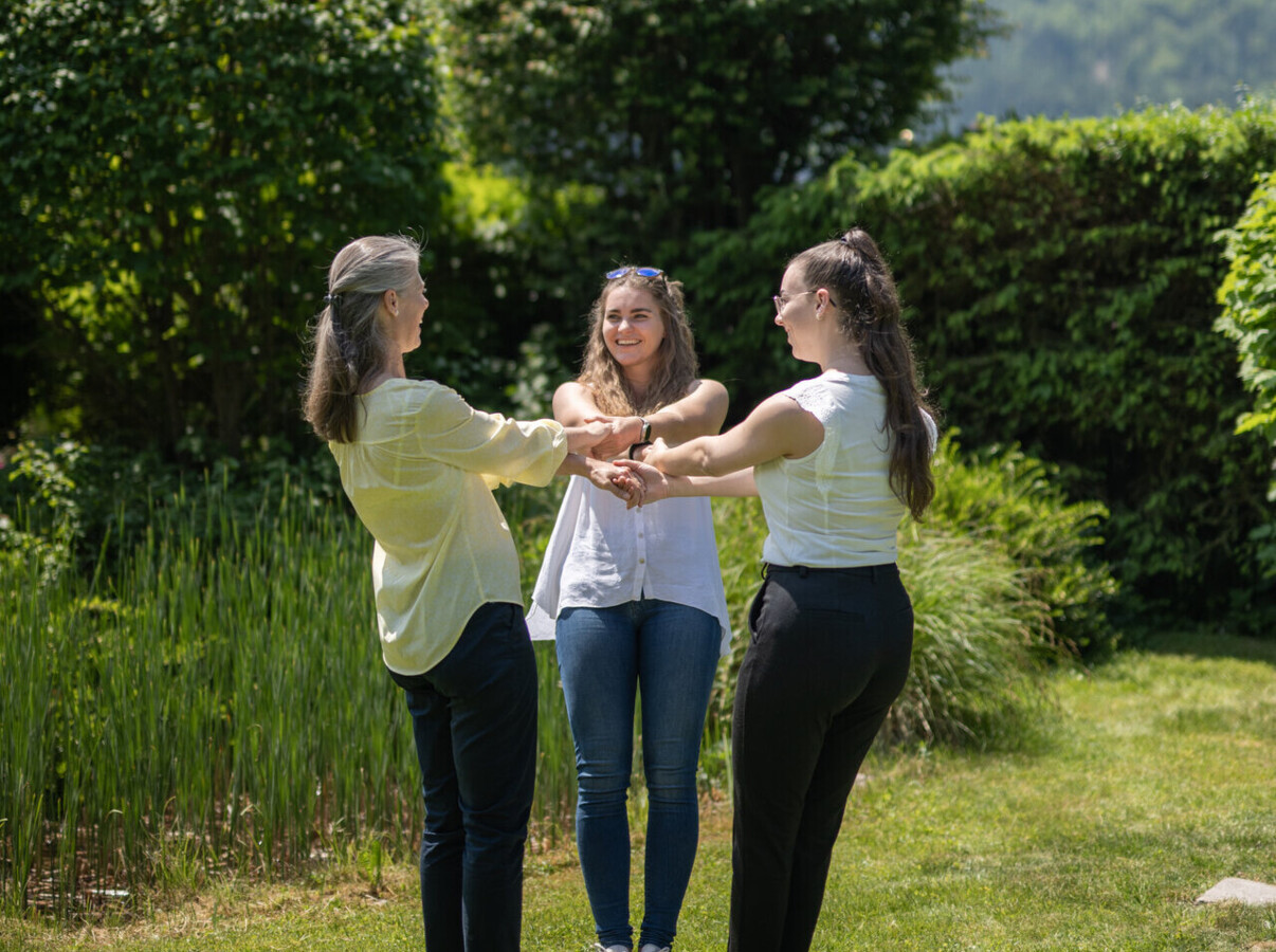 Drei Damen stehen im Kreis und halten sich an den Händen im Garten des Seminarhotel Waldhof Fuschlsee Resort.