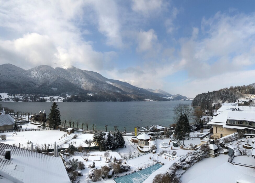Snow-covered landscape with lake Fuschl and mountains in the background, surrounded by buildings and trees under a partly cloudy sky.