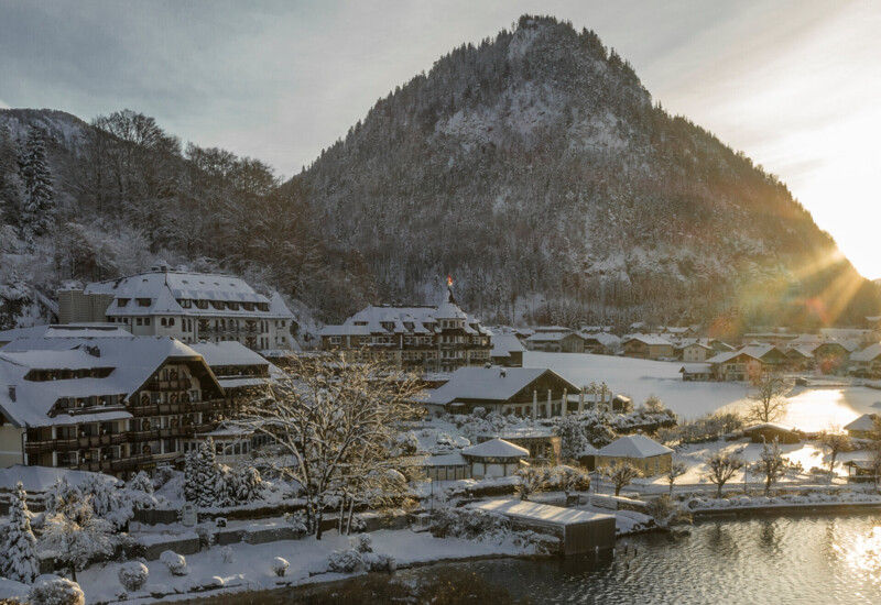 Snow-covered village by lake Fuschl with a mountain backdrop and sunlight streaming through trees.