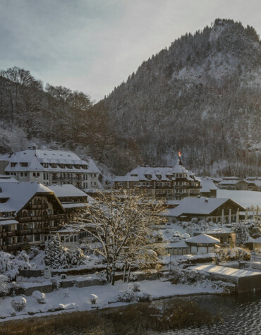 Snow-covered village by lake Fuschl with a mountain backdrop and sunlight streaming through trees.