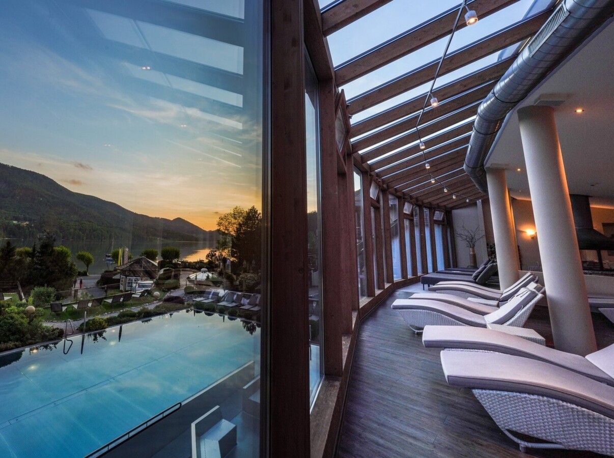 Glass-walled lounge with sun chairs overlooking a scenic lake and mountains at sunset.