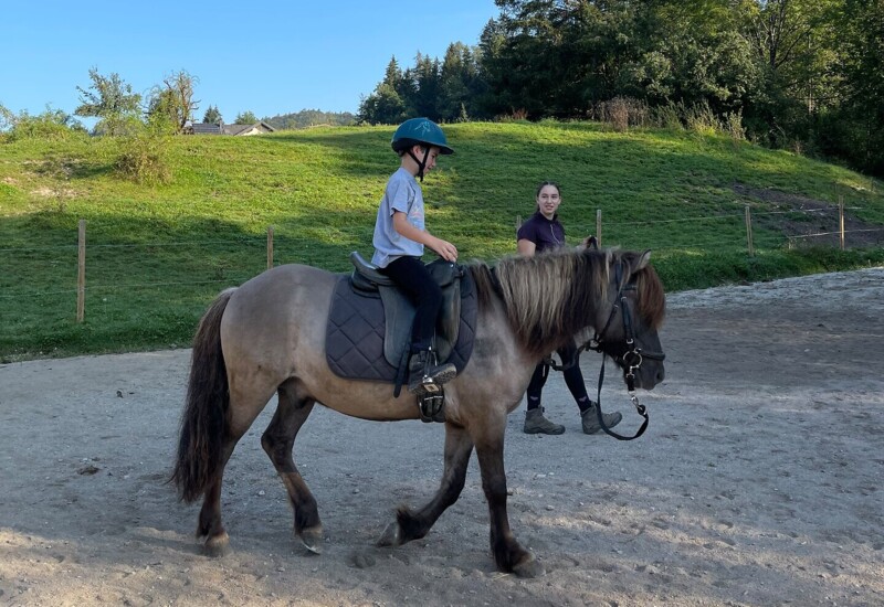 Kind reitet auf einem Pferd in grüner Landschaft unter blauem Himmel, begleitet von einem Erwachsenen.