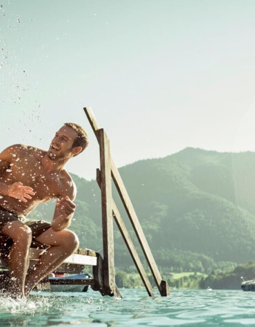 Ein Paar sitzt lachend auf einem Steg am Fuschlsee, während Wasser spritzt. Im Hintergrund sind Berge und ein Segelboot zu sehen.