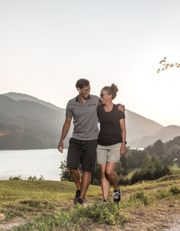 Ein Paar spaziert bei Sonnenuntergang am Fuschlsee entlang, umgeben von grünen Hügeln und ruhiger Natur.