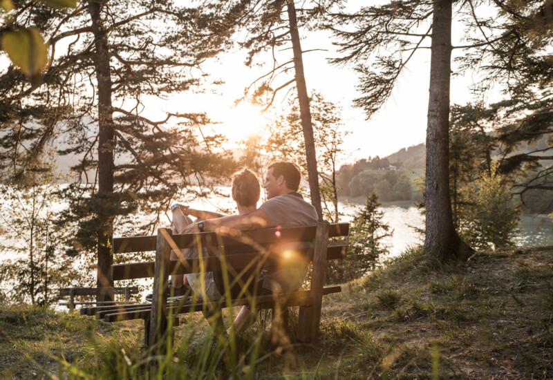 Ein Paar sitzt auf einer Bank im Wald und genießt den Sonnenuntergang am Fuschlsee.